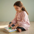 SuYa Shop pink gingham school smock with elastic cuffs worn by a girl sitting on the floor reading a book